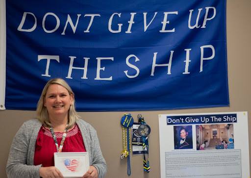 Betsy Allee stands in front of a U.S. Navy flag and other Navy souvenirs while holding a drawing from one of her students. The flag is a replica of the original, which hangs in Memorial Hall in the U.S. Naval Academy (USNA) and stands to commemorate former graduates who have died in service.