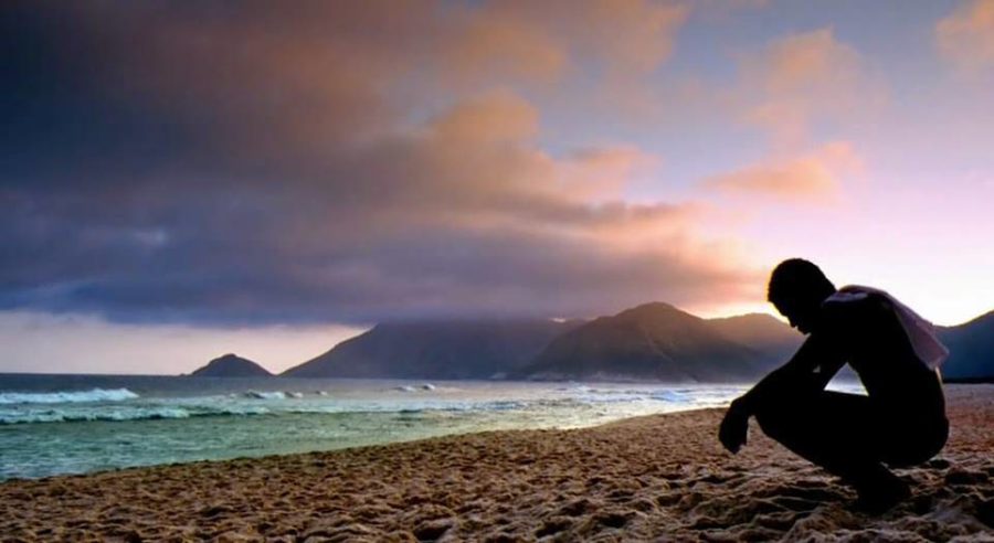 Rocket (played by Alexandre Rodrigues) sits pensively on a beach as the sun sets over the sea