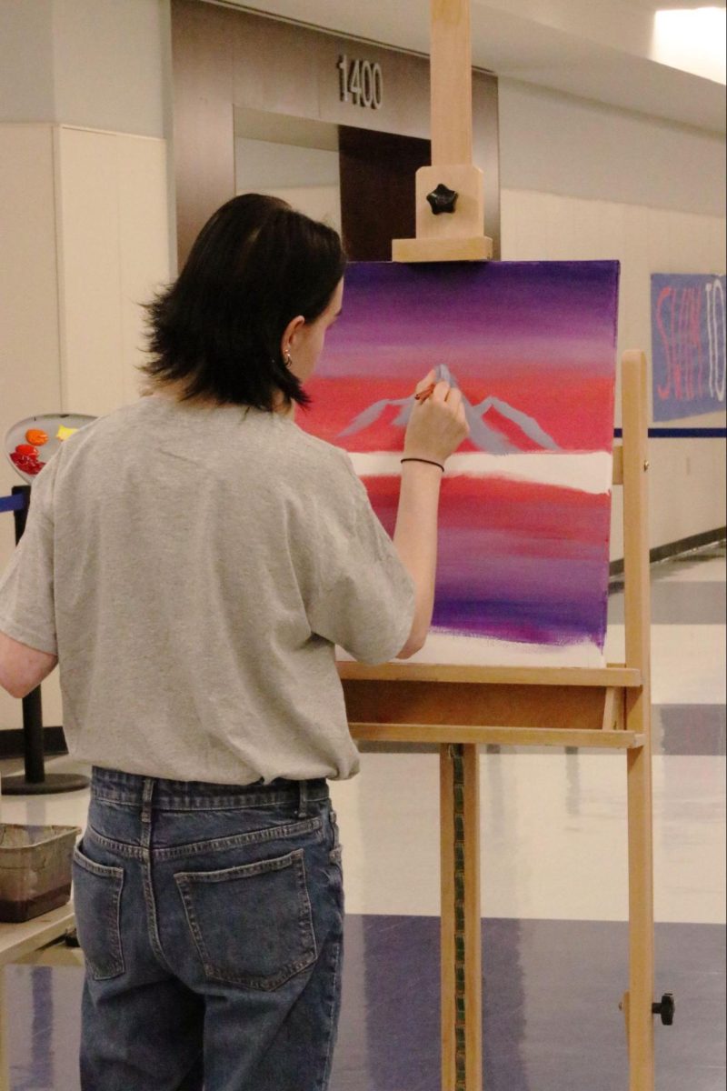 Senior Asher Hull paints a mountain over a lake with trees during sunset. This was all made live during the Art Gallery Showing.